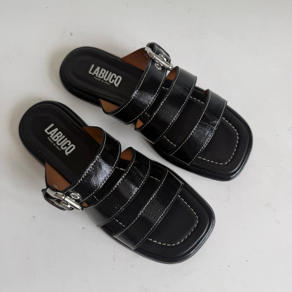 A pair of black leather slide sandals with double straps and silver buckle details, displayed on a white surface against a white wall.