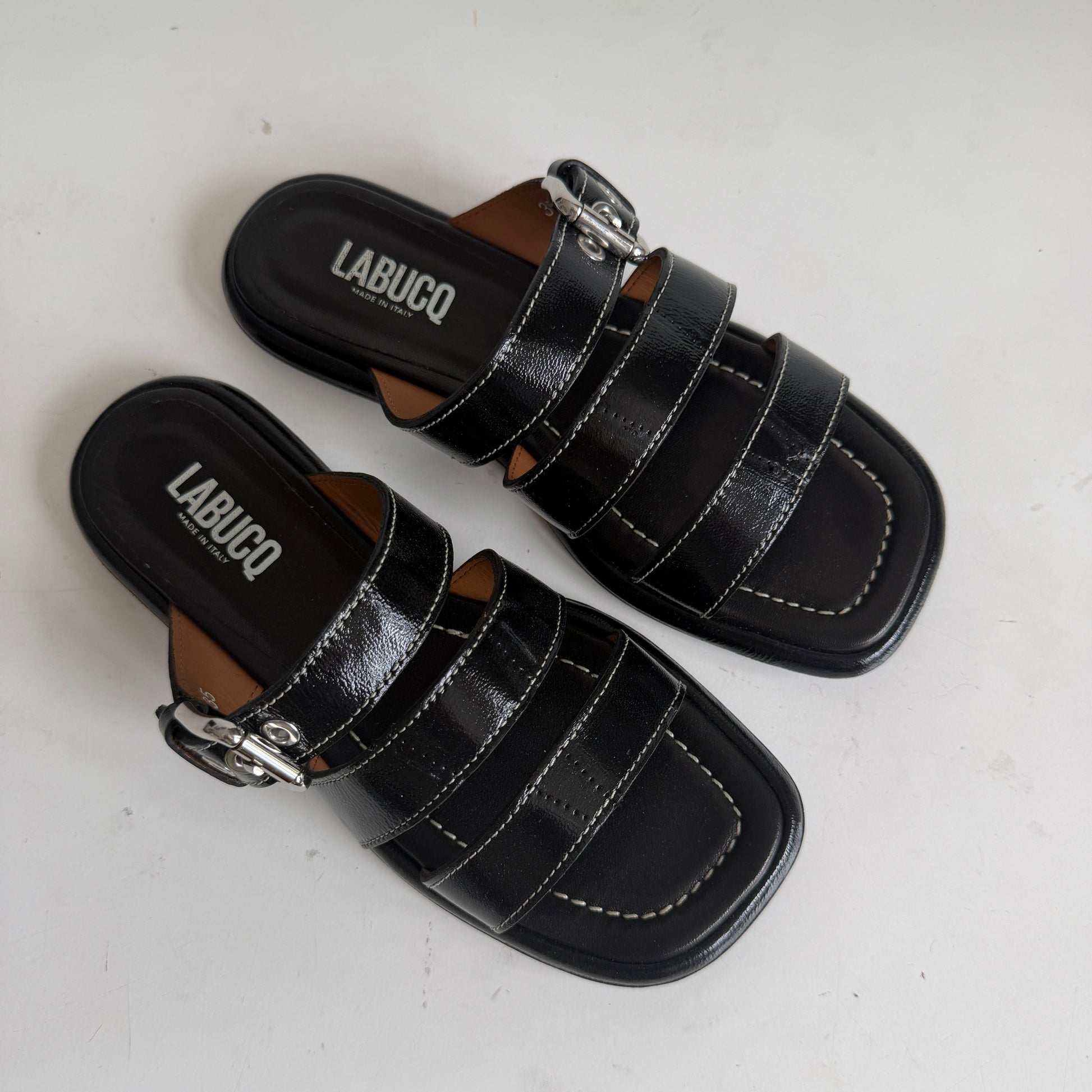A pair of black leather slide sandals with double straps and silver buckle details, displayed on a white surface against a white wall.