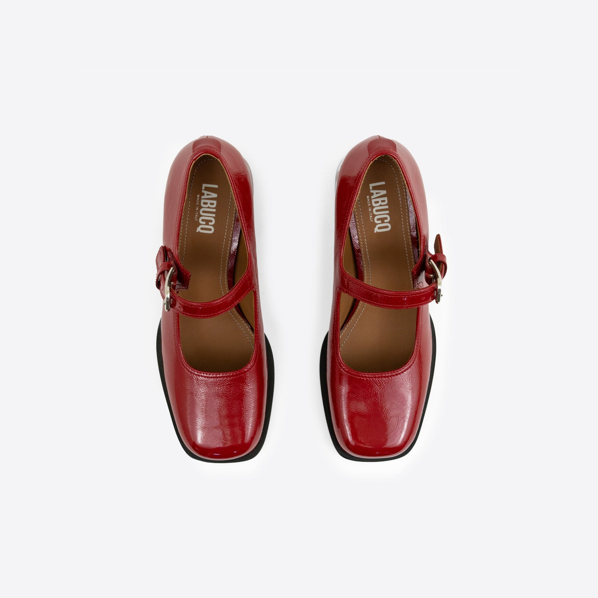 Top-view shot of a pair of red patent leather Maryjanes with visible Labucq branding, a buckle, and a black sole on a white background
